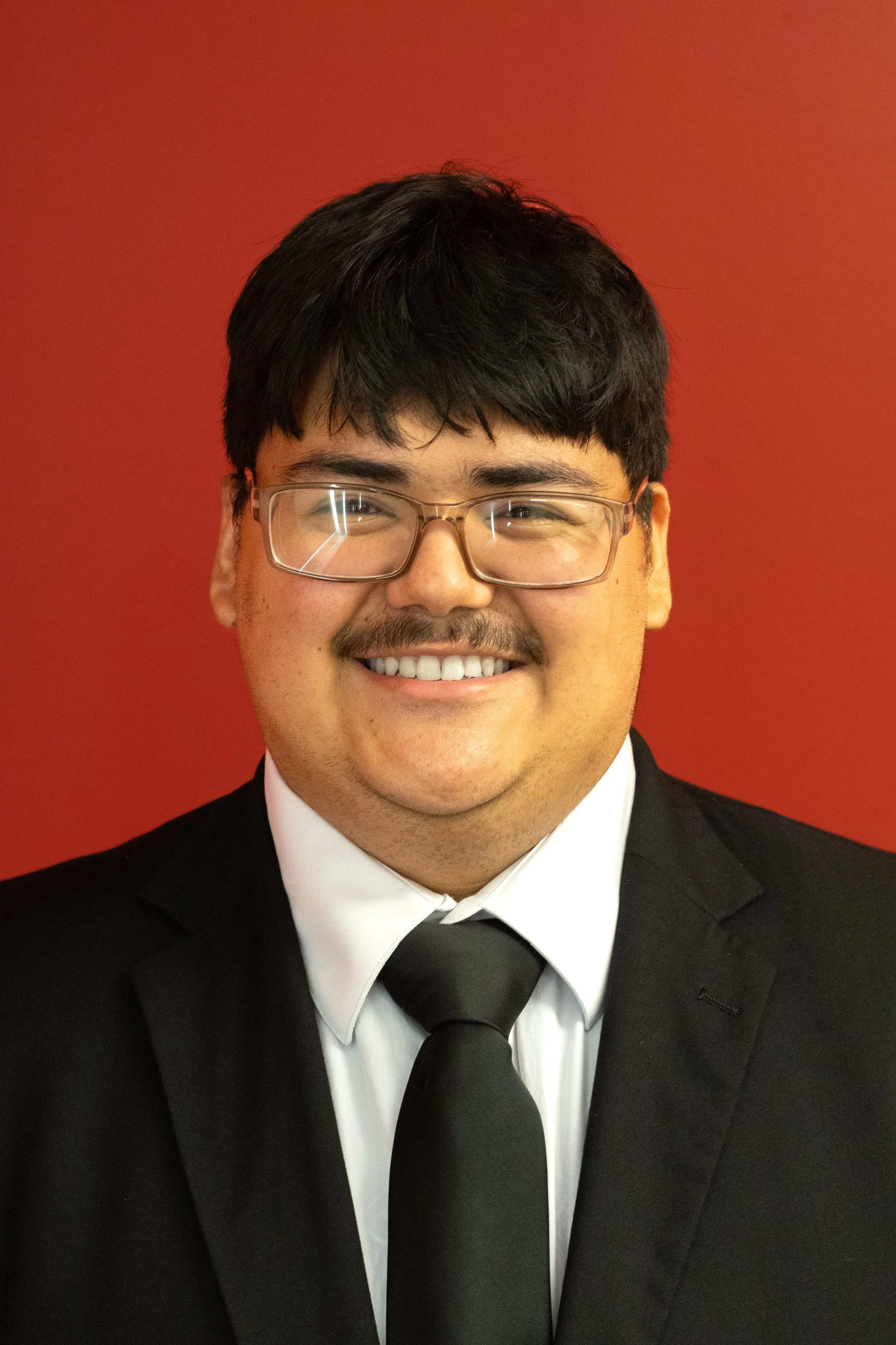 A man with glasses, a mustache, and dark hair is smiling at the camera. He is wearing a suit and tie with a white shirt, set against a red background.