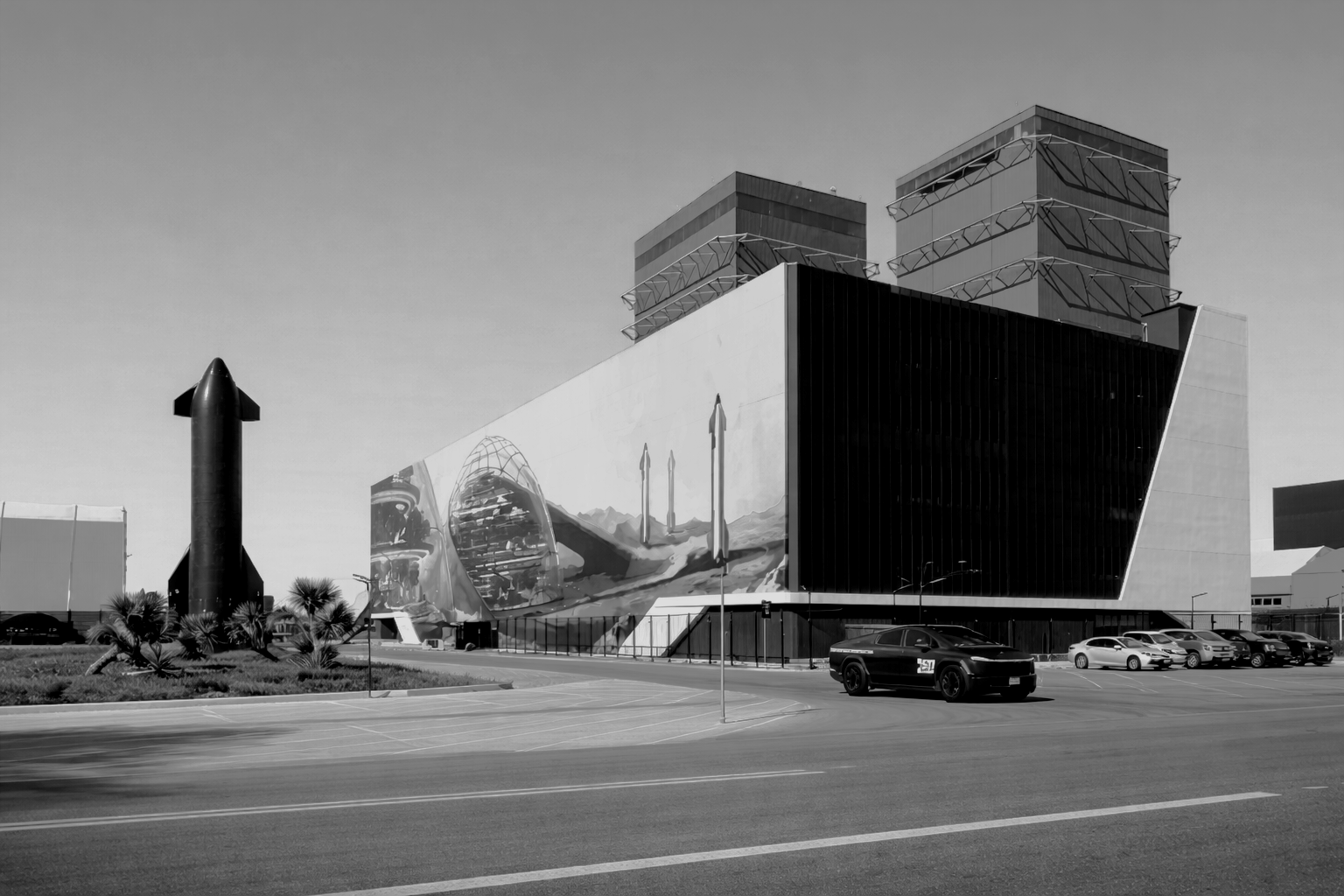 Black and white photo of a modern building with large mural, two tall structures, a parking lot with cars, and a rocket sculpture in the foreground.