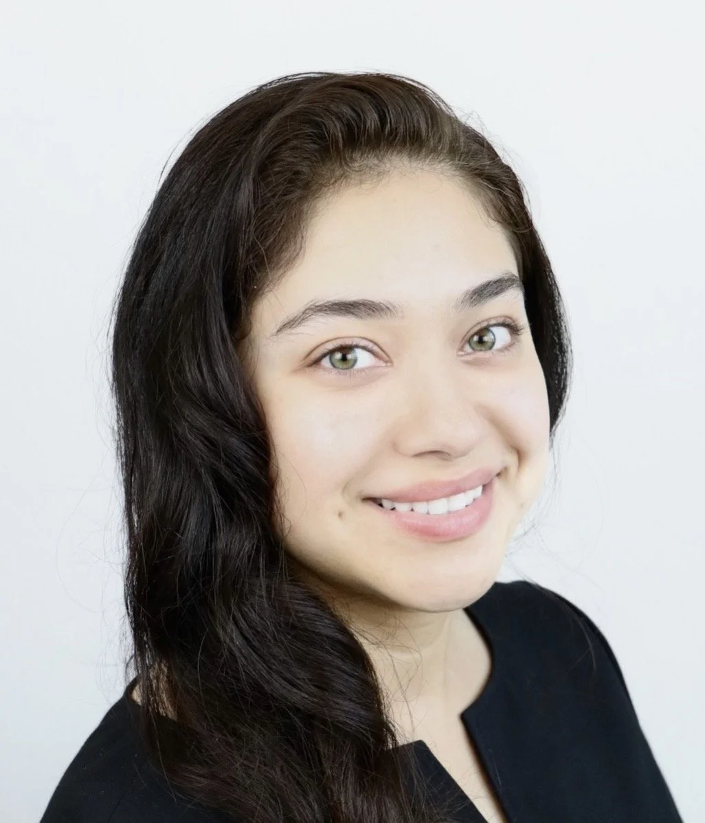 Close-up of a smiling young woman with long dark hair, fair skin, and light-colored eyes, wearing a black top.