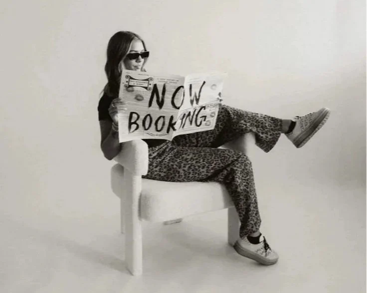 Woman sitting in a chair, wearing sunglasses and casual clothes, holding a sign that reads 'NOW BOOKING'.
