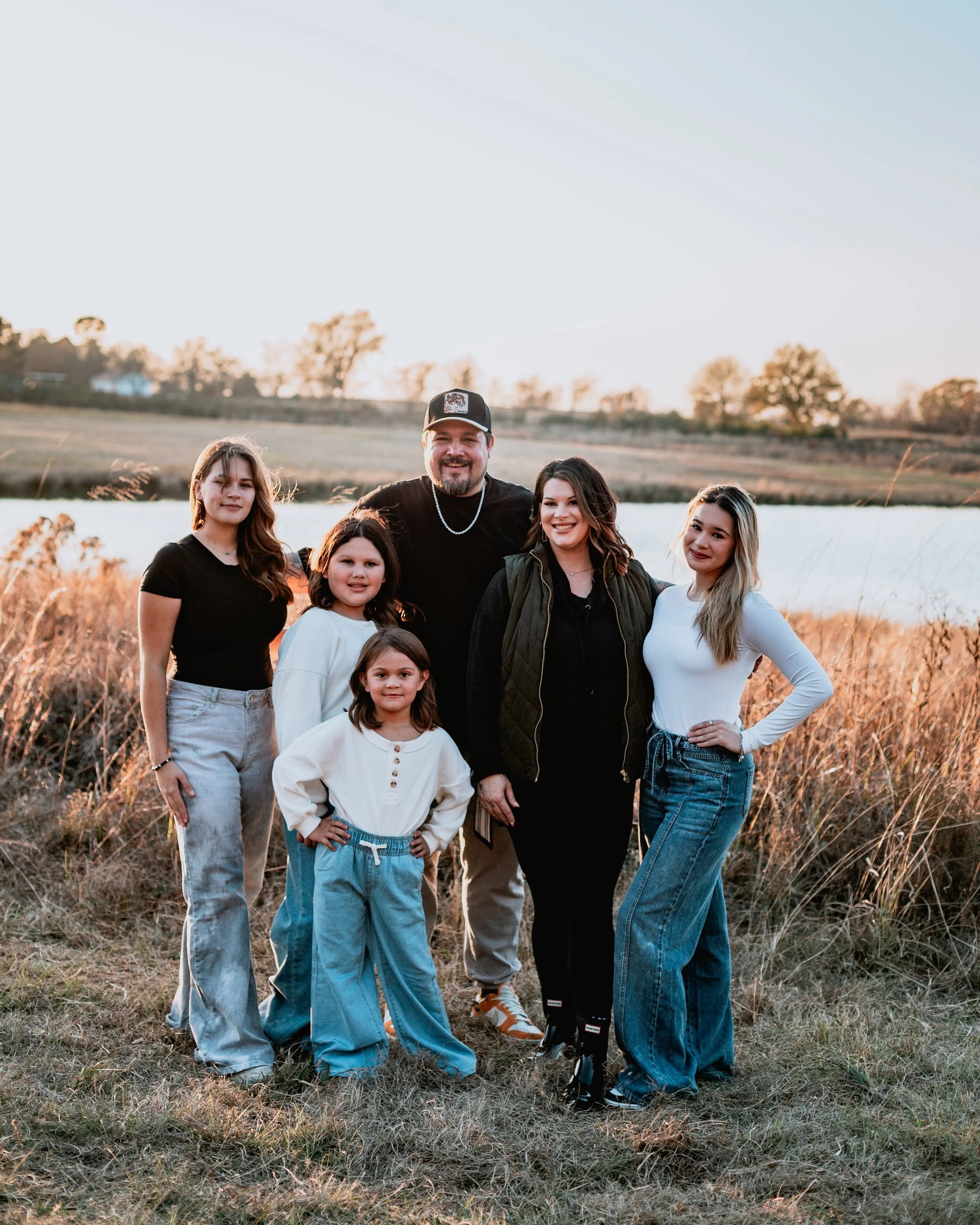 A family of six posing outdoors near a body of water during sunset. The group includes two adults and four children, standing on grassy terrain with dry bushes and trees in the background.