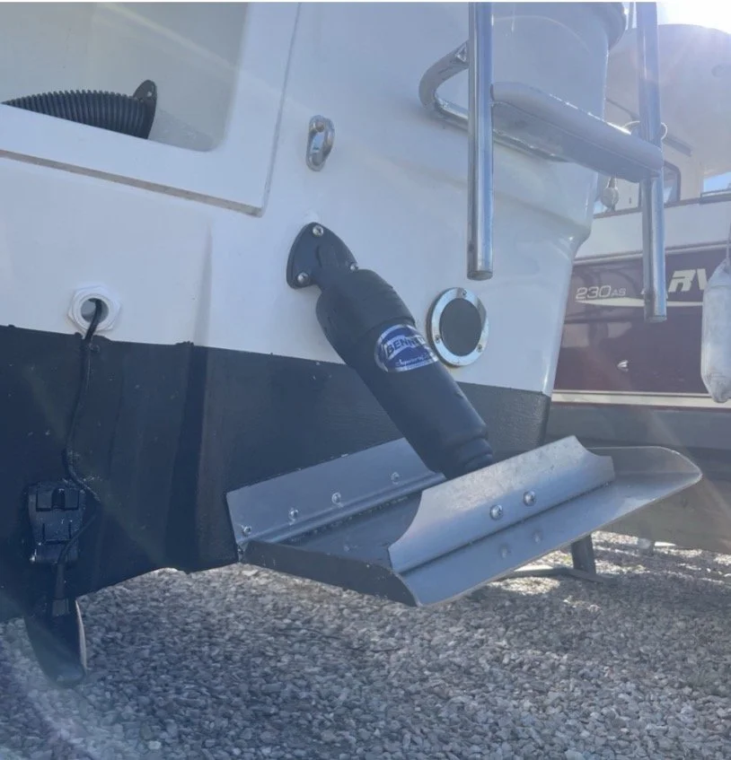 Close-up of the bow of a boat showing an underwater thruster attached at the bottom, with a black and blue cover and a metal skid plate underneath. A part of an engine or propeller is visible on the right.