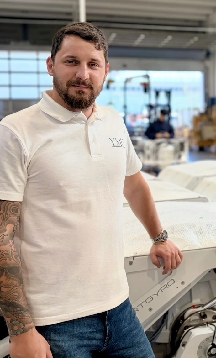 A man with a beard and tattoos on his left arm, wearing a white polo shirt and blue jeans, standing inside a large industrial or manufacturing building. He is leaning on a piece of machinery, with a person working in the background.