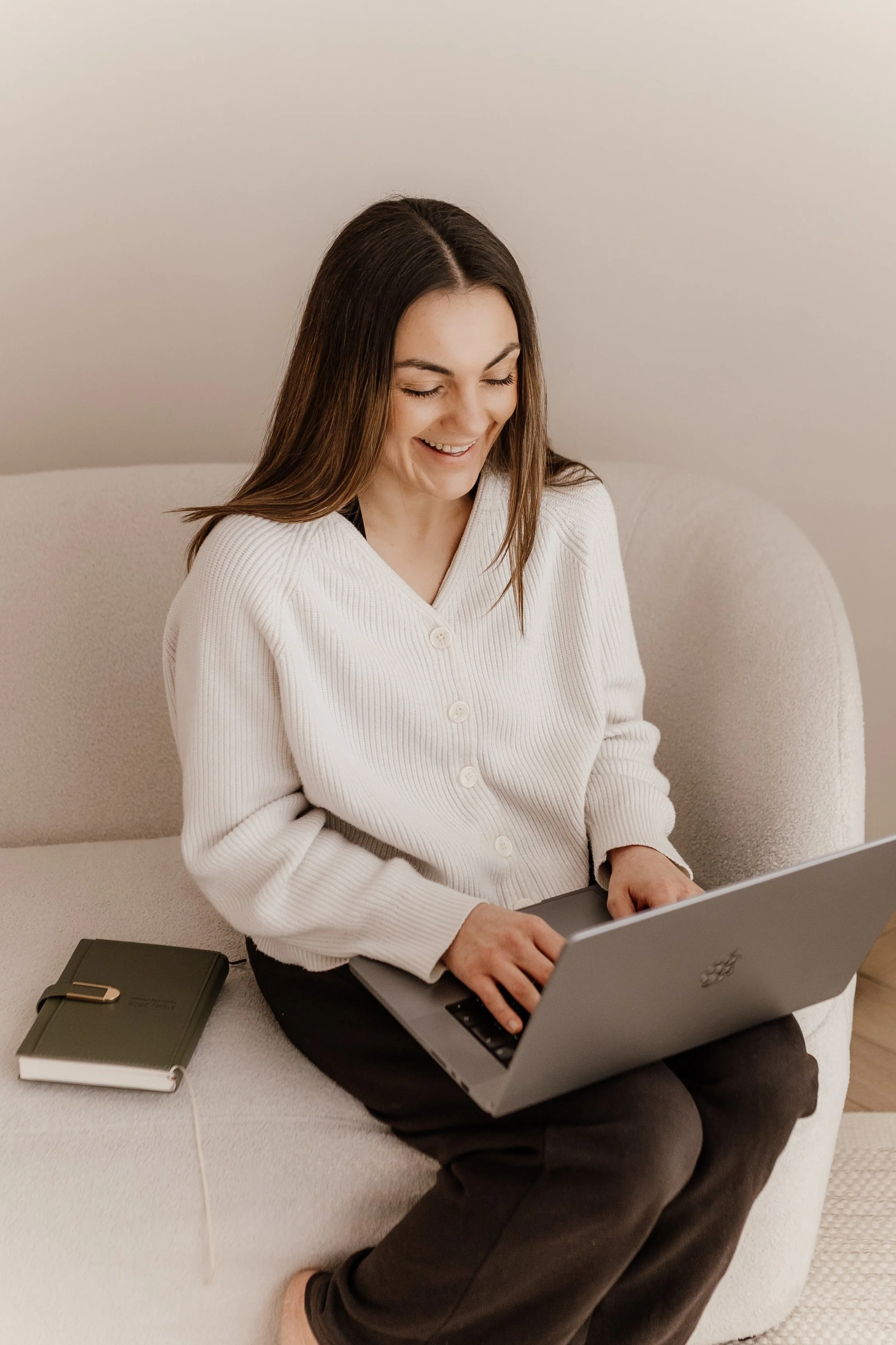 A woman sitting on a beige sofa, typing on a silver laptop, smiling, with a closed notebook beside her.