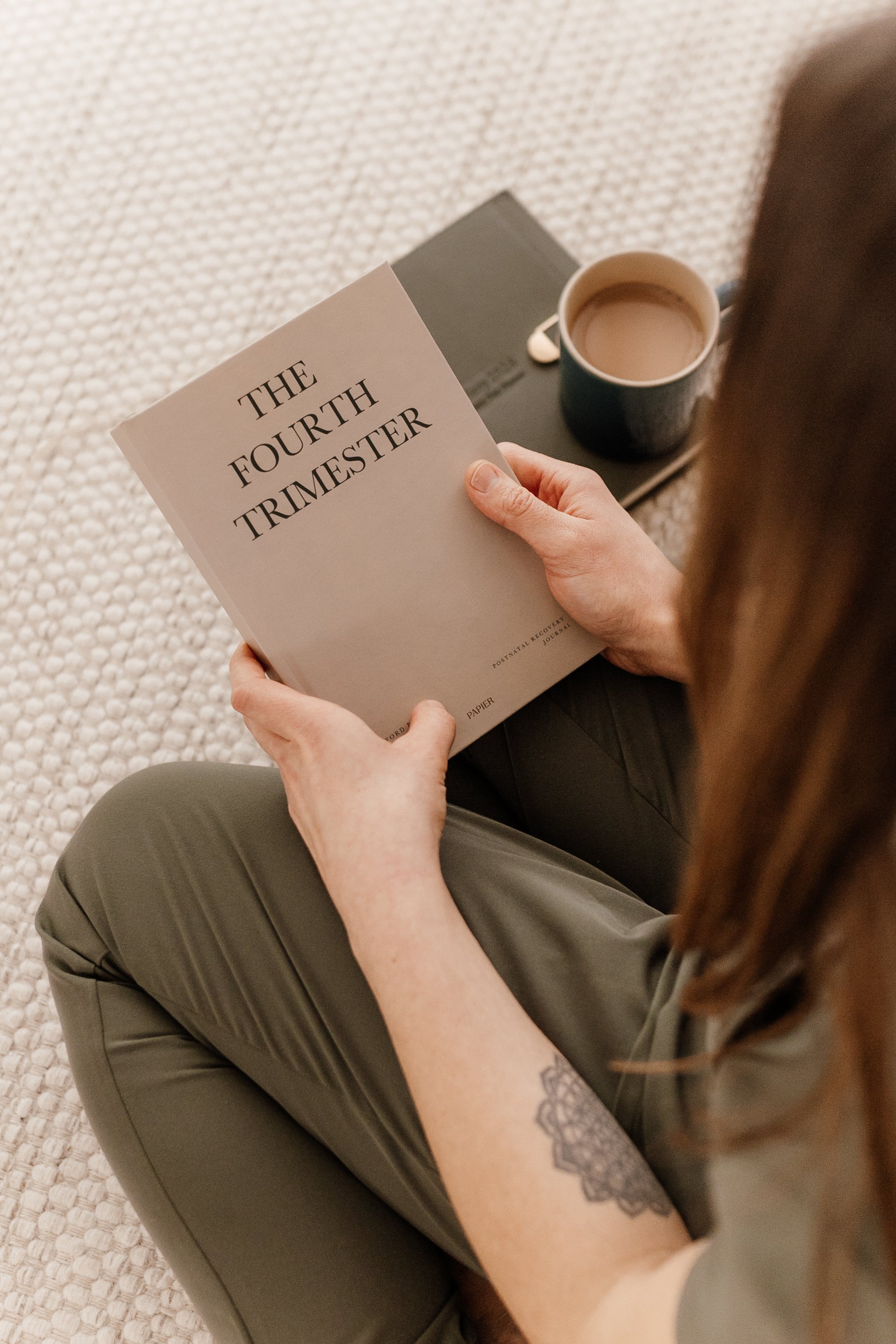 A person sitting on a beige textured carpet, holding a book titled 'The Fourth Trimester' with a coffee mug and a closed laptop on the floor nearby.