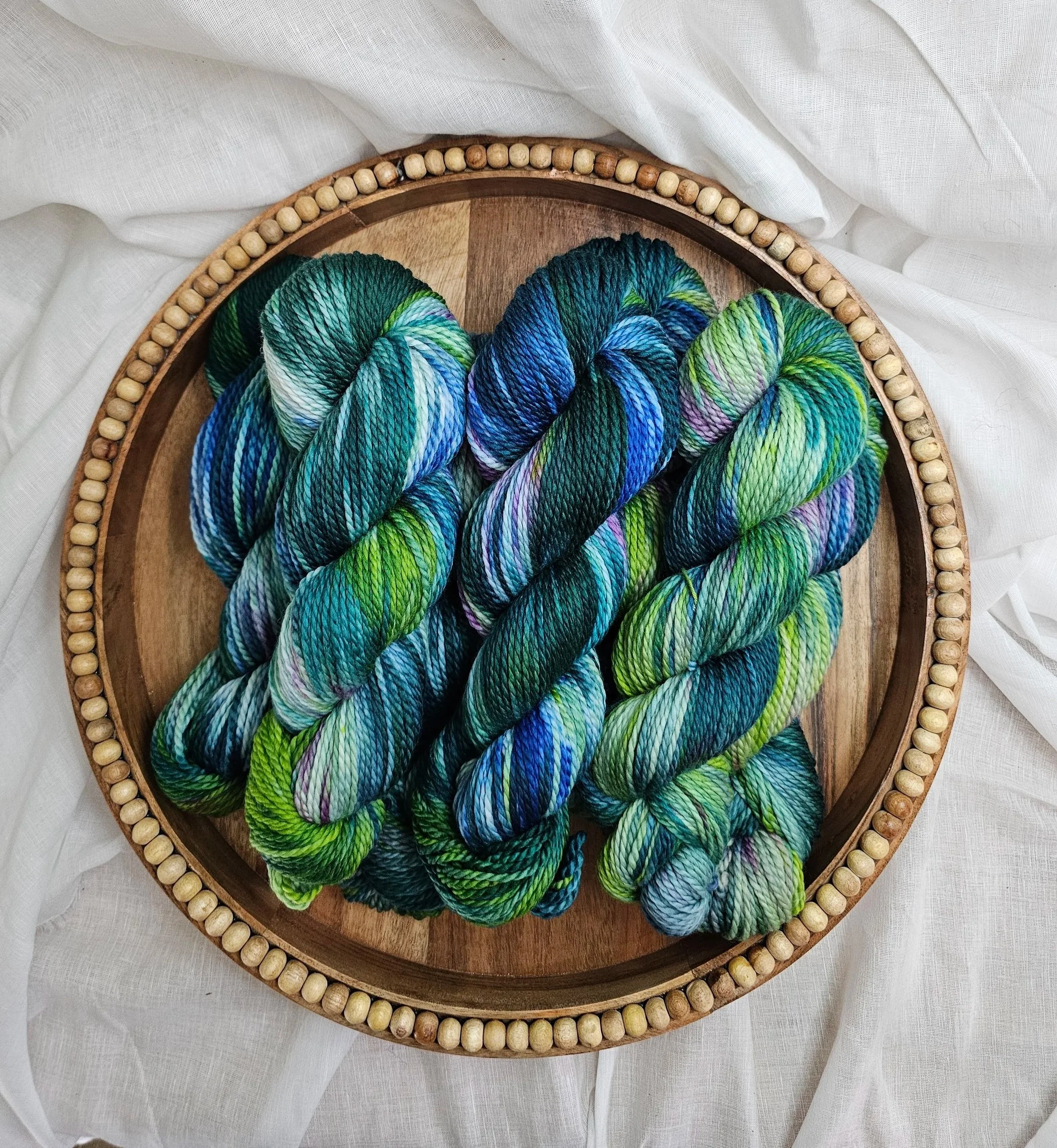 Colorful skeins of yarn in shades of blue, green, purple, and black inside a round wooden embroidery hoop, on a white fabric background.