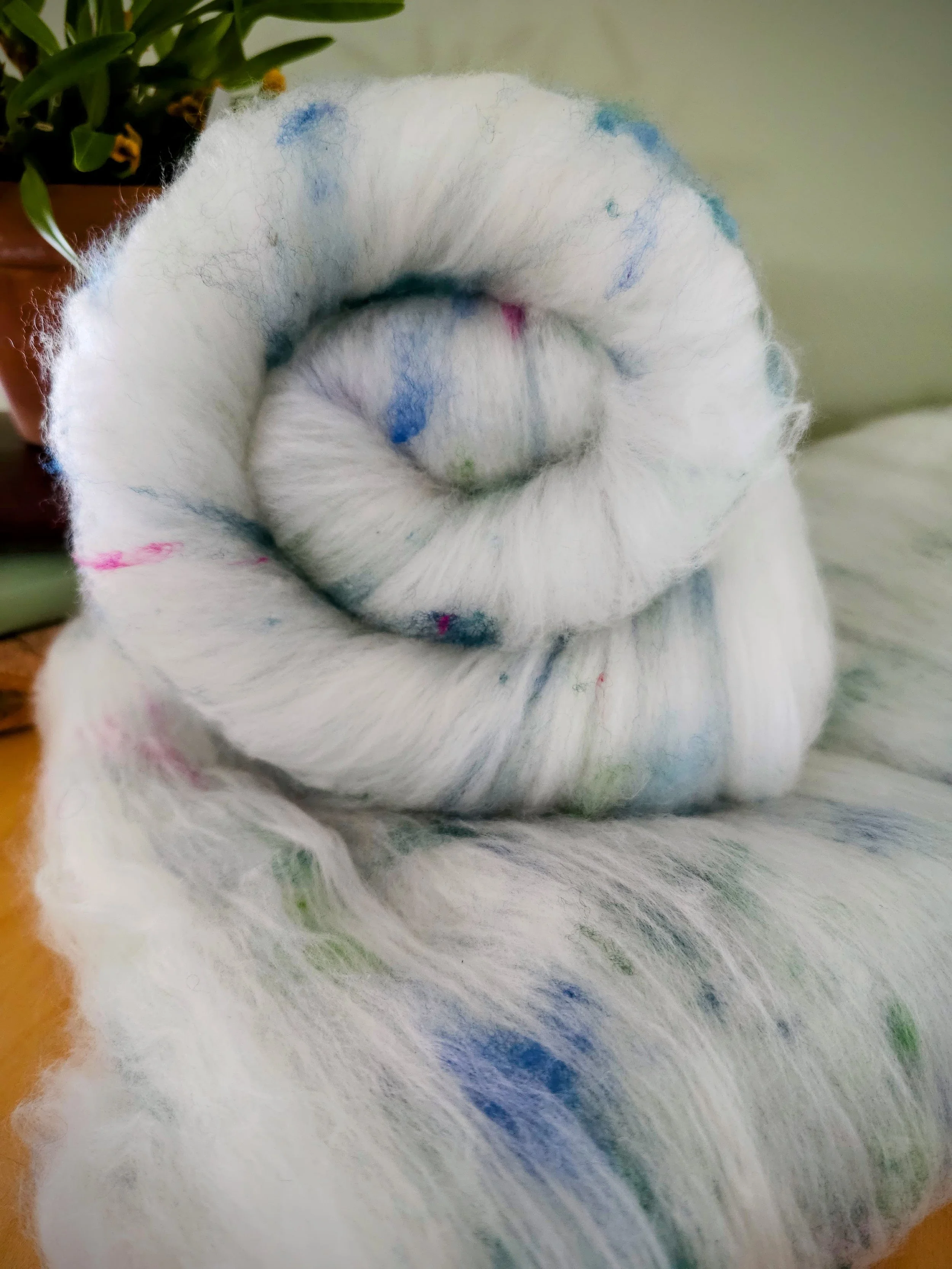 Close-up of a white wool batt with colorful pops of blue and green splatters, draped over a surface near a potted plant.