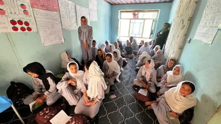 School classroom with young girls in gray uniforms and white headscarves sitting on the floor, some with notebooks, while teachers stand near the window and walls.