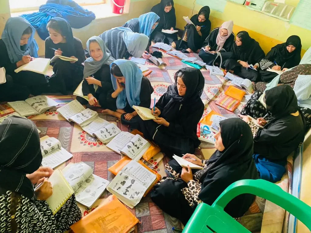 A group of women and girls sitting on the floor of a classroom, reading and taking notes from open books. Some are wearing hijabs and traditional clothing, with educational materials scattered around.