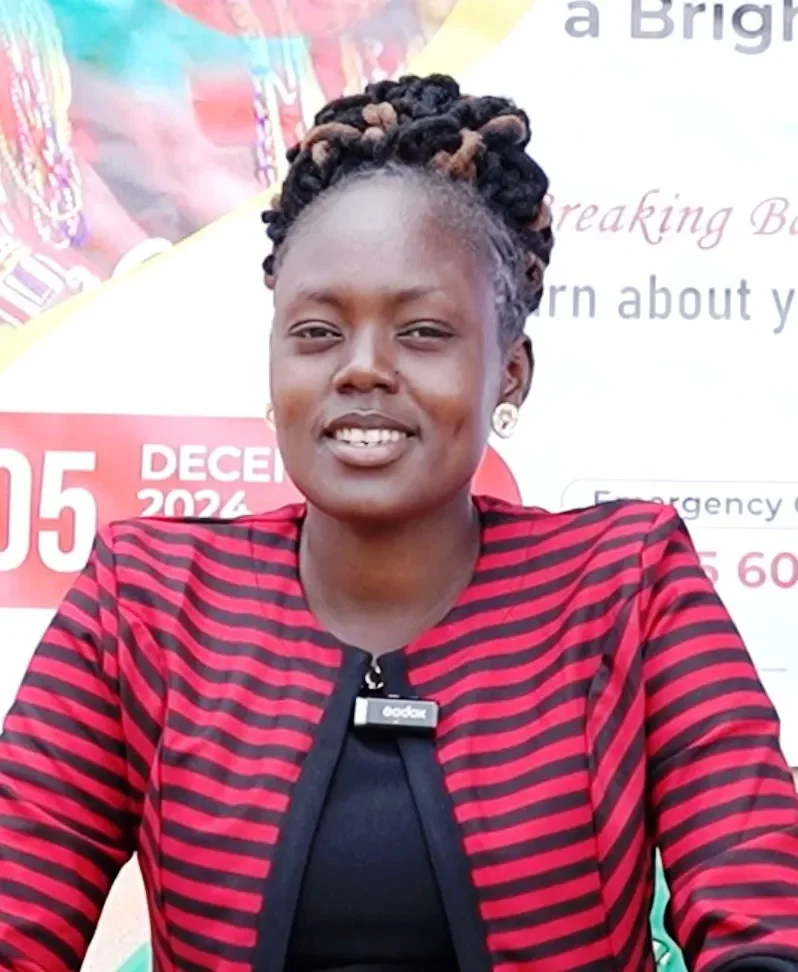A woman with braided hair wearing a red and black striped jacket, sitting in front of a promotional banner.