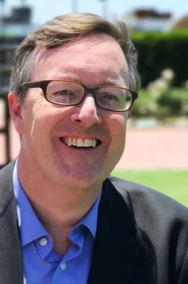 Smiling man with glasses outdoors, wearing a blue shirt and dark blazer.
