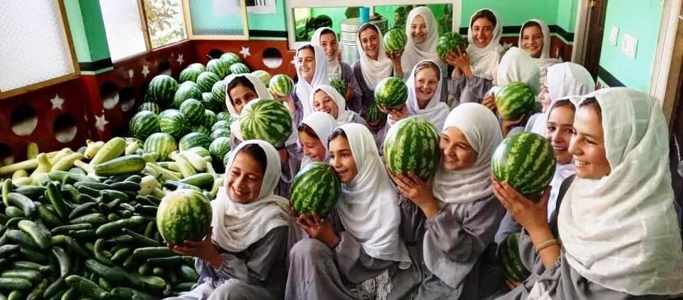 Young girls in school uniforms with white headscarves holding watermelons, smiling in a classroom surrounded by piles of watermelons and cucumbers.