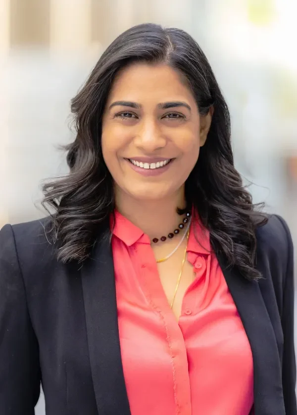 A woman with dark wavy hair, smiling, wearing a coral blouse and a navy blazer, standing outdoors.