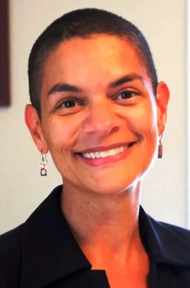 Smiling woman with short dark hair, earrings, wearing a black top, against a light-colored background.