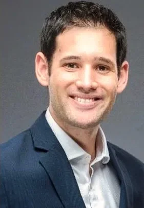 A young man with dark hair and light skin, wearing a navy blazer and white shirt, smiling at the camera against a gray background.