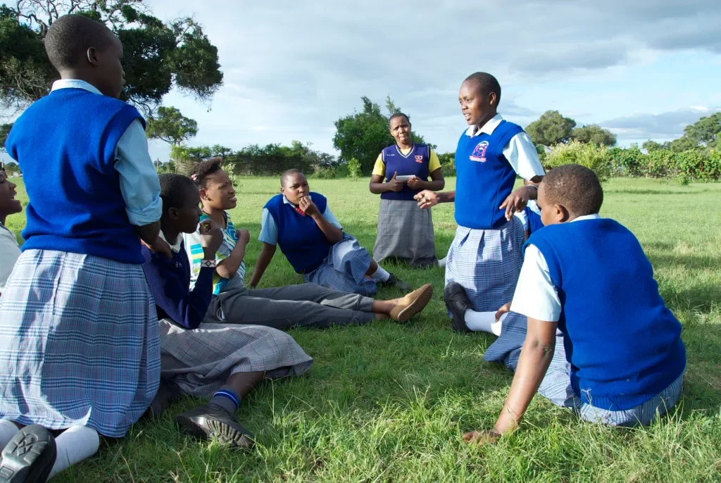 A group of students in blue school uniforms sitting and listening to a girl in the same uniform standing and speaking outdoors on a grassy field.