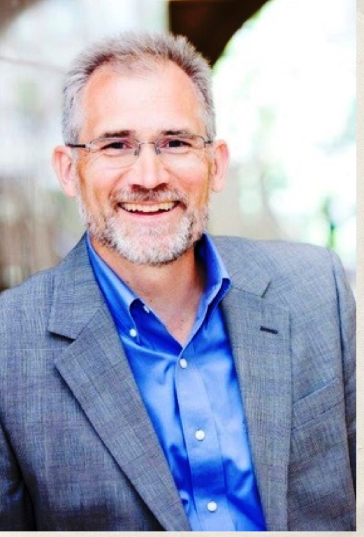 A middle-aged man with gray hair and a beard, wearing glasses, a blue shirt, and a gray blazer smiling.