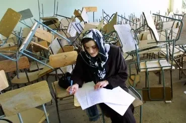A woman sitting in a classroom filled with overturned and displaced chairs, studying from an open book.