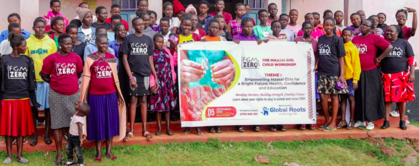 A group of women and girls standing outdoors in front of a building, holding a banner promoting a girls' workshop on health, confidence, and education. The banner indicates it is organized by FGM Zero and sponsored by Global Roots. Some women are wearing shirts with FGM Zero written on them, and children are present among the group.