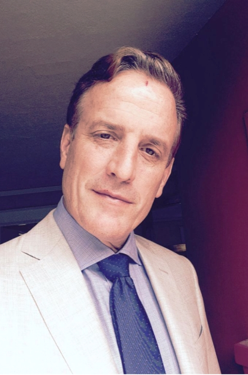 A man in a beige suit, light blue shirt, and dark blue tie smiling for the camera indoors.