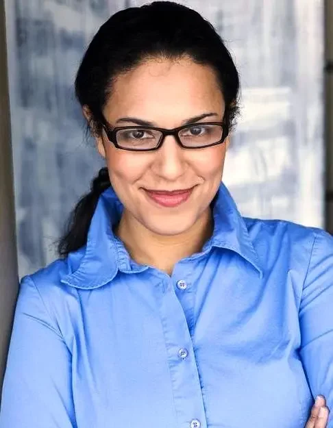 A woman with dark hair in a ponytail, wearing glasses and a blue button-up shirt, standing in front of a window with blinds, smiling and crossing her arms.
