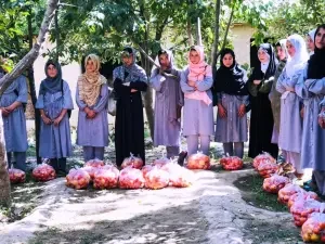 Group of women and men standing outdoors in a circle around flower-covered graves in a cemetery, engaging in a memorial or prayer gathering.