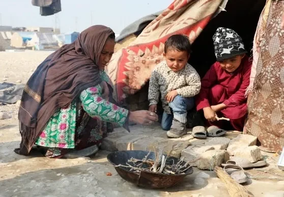 A woman and two children sitting on the ground next to a makeshift tent, with shoes nearby, in a desert-like setting.