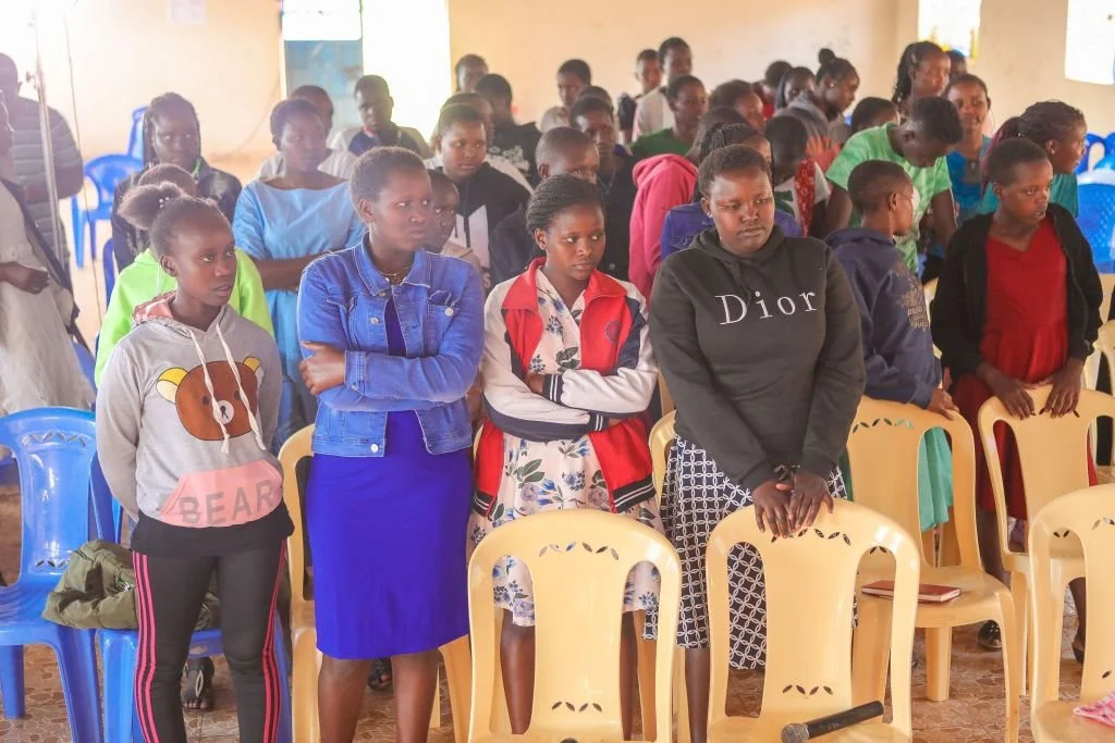 Group of children standing in a classroom, some with arms crossed, facing forward.