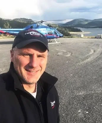 A man smiling in front of a helicopter near a body of water with mountains in the background.