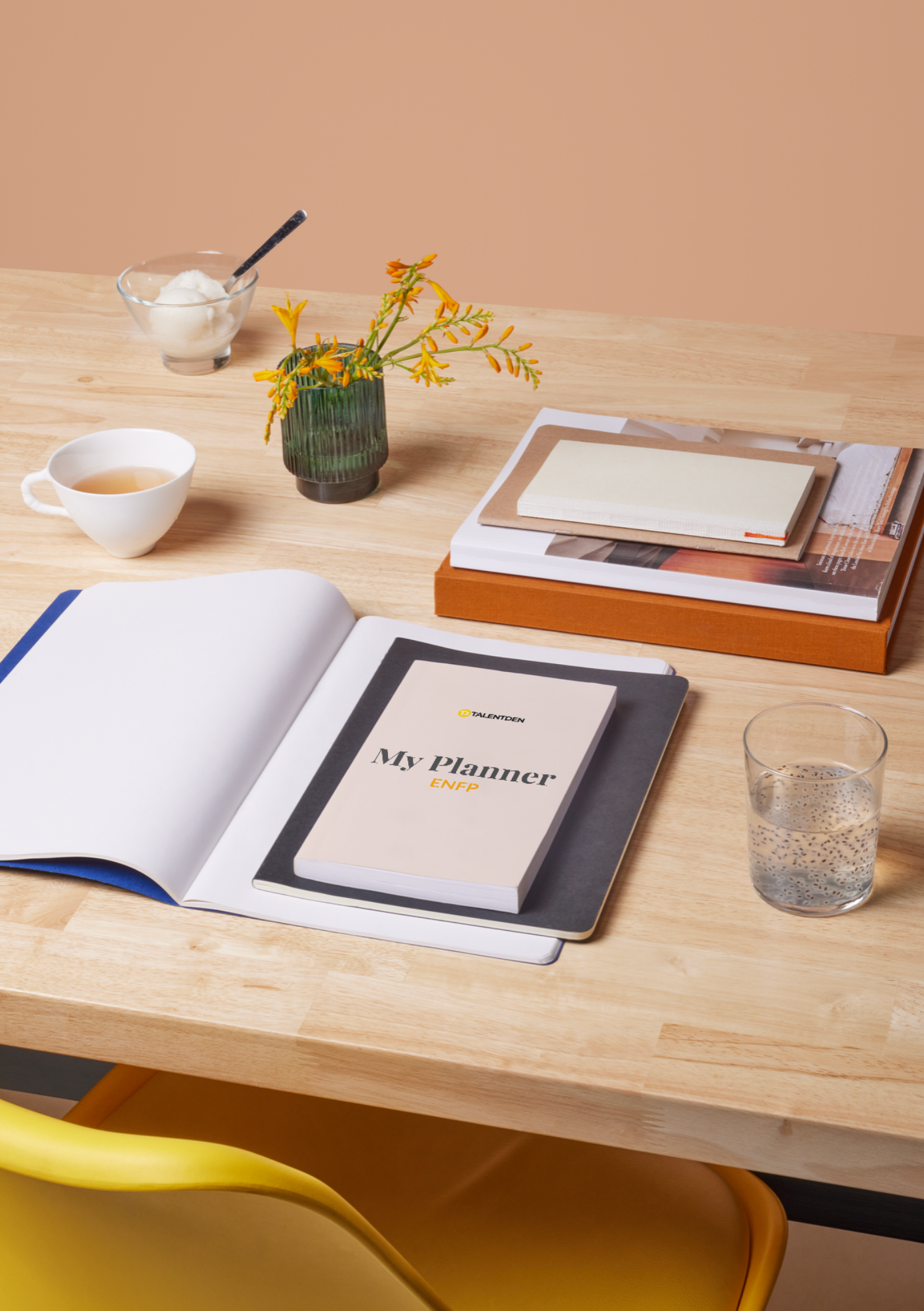Open planner on wooden table with a book titled 'My Planner ENFP', a glass of water, a notebook, a small white book, a vase with yellow-orange flowers, a cup of tea, a glass of water with chia seeds, and a bowl of ice cream with a spoon.