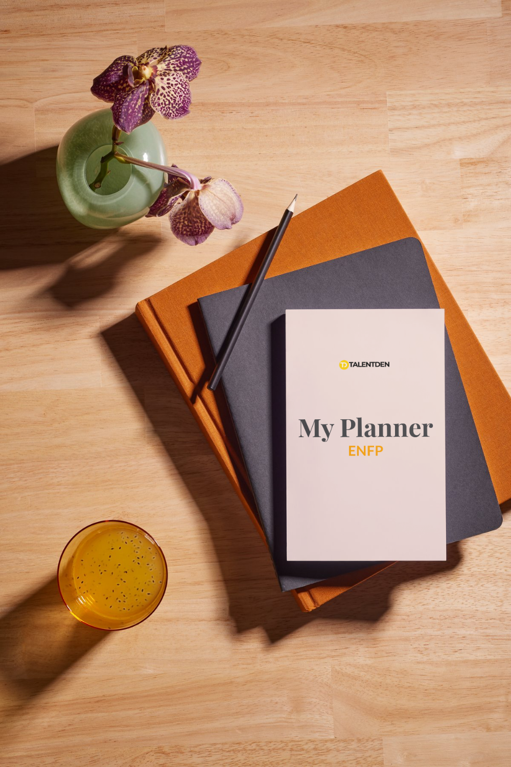 A top-down view of a wooden desk with a potted orchid, a yellow drink in a glass, and stacked notebooks with a planner titled 'My Planner ENFP' on top.