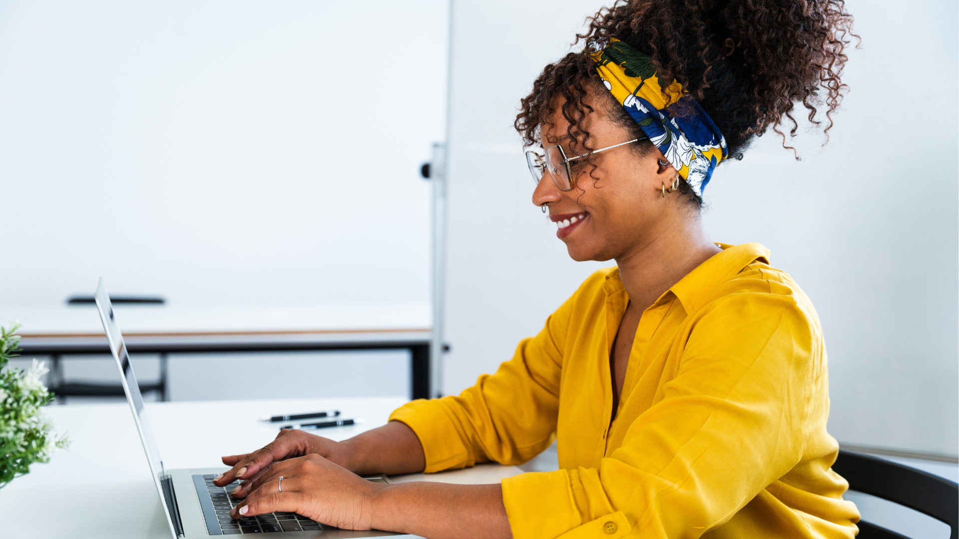 Woman with curly hair, glasses, and gold earrings, wearing a yellow shirt and a colorful headband, working on a laptop at a desk.