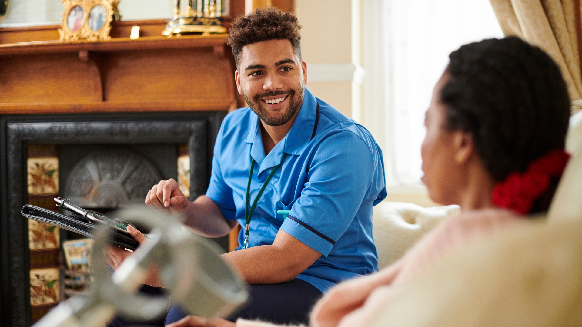 A young man in a blue uniform smiling and talking to a woman sitting in a chair in a cozy room with a fireplace and decorative items.