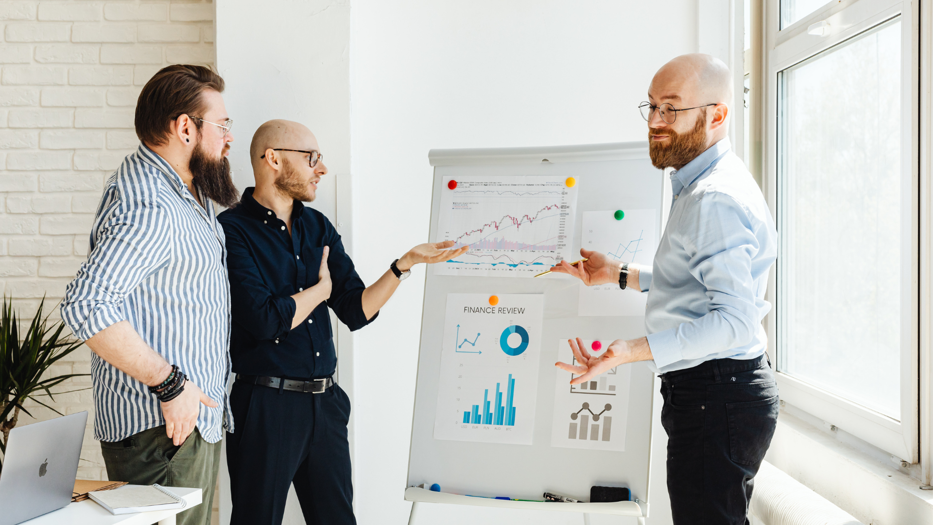 Three men in business casual attire discussing financial graphs and data on a presentation board in a well-lit office.