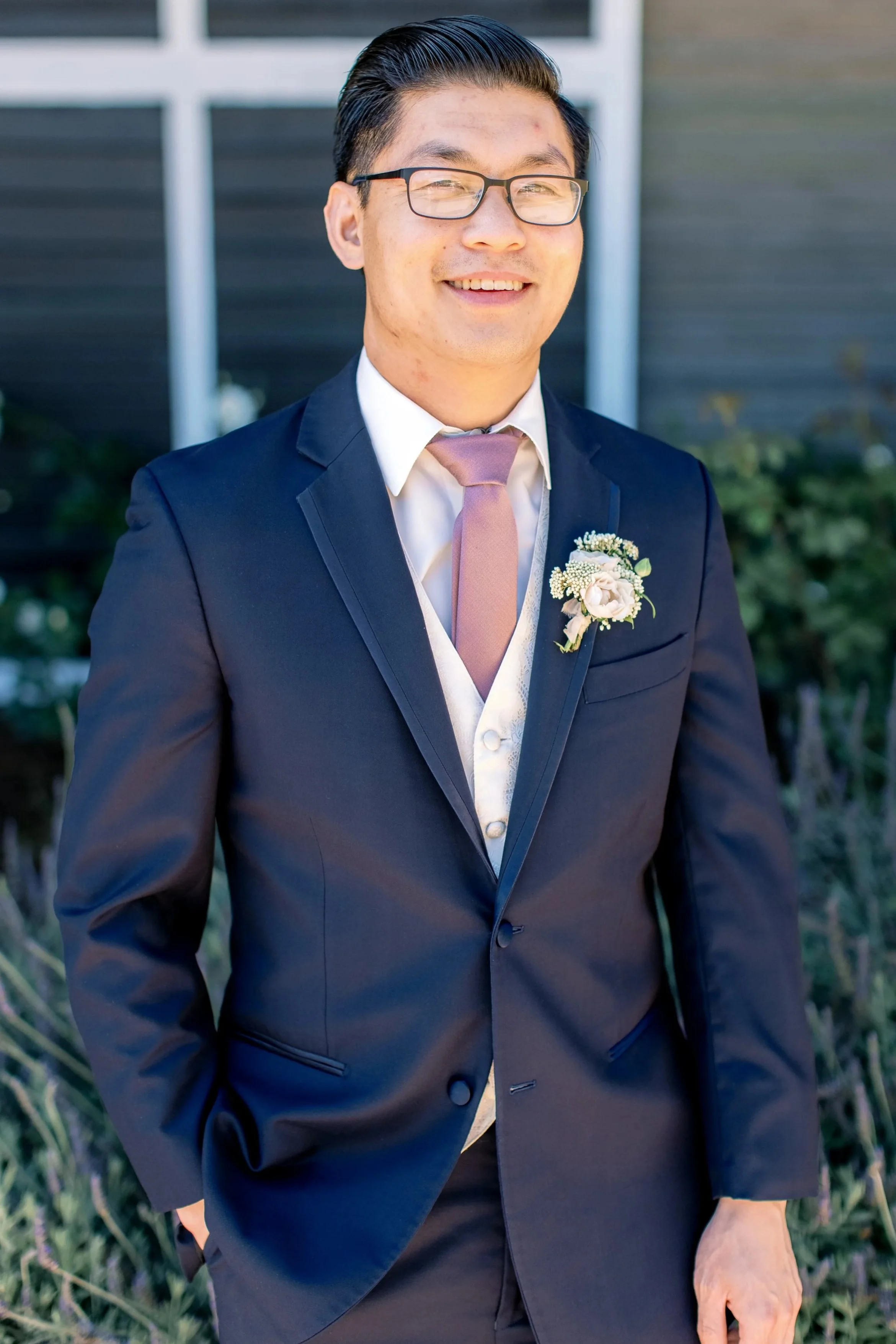 Smiling man in glasses wearing a navy suit with a boutonniere, white shirt, and pink tie outdoors.