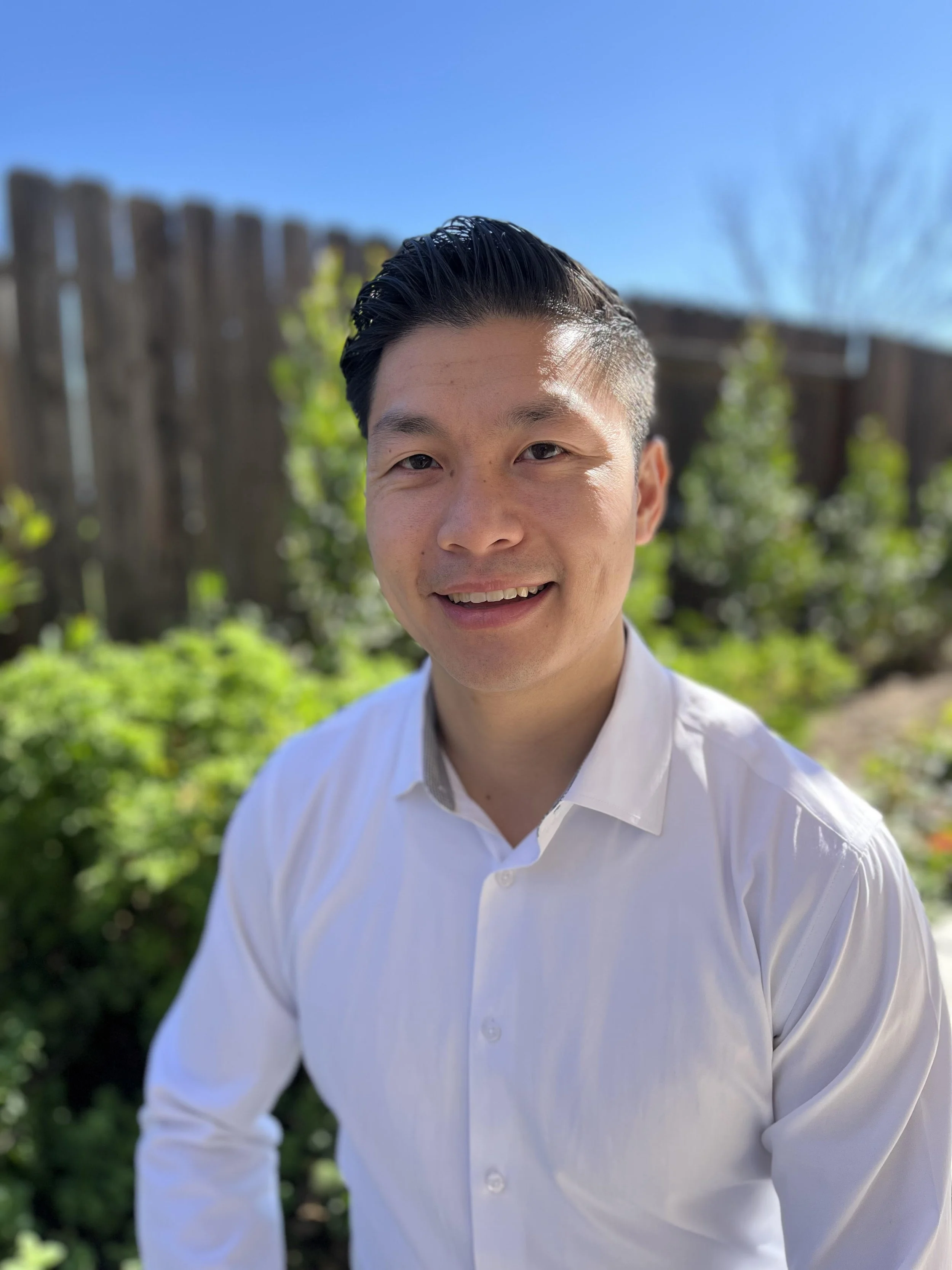 A young man with dark, slicked-back hair wearing a white dress shirt, smiling outdoors with a wooden fence and greenery in the background under a bright blue sky.