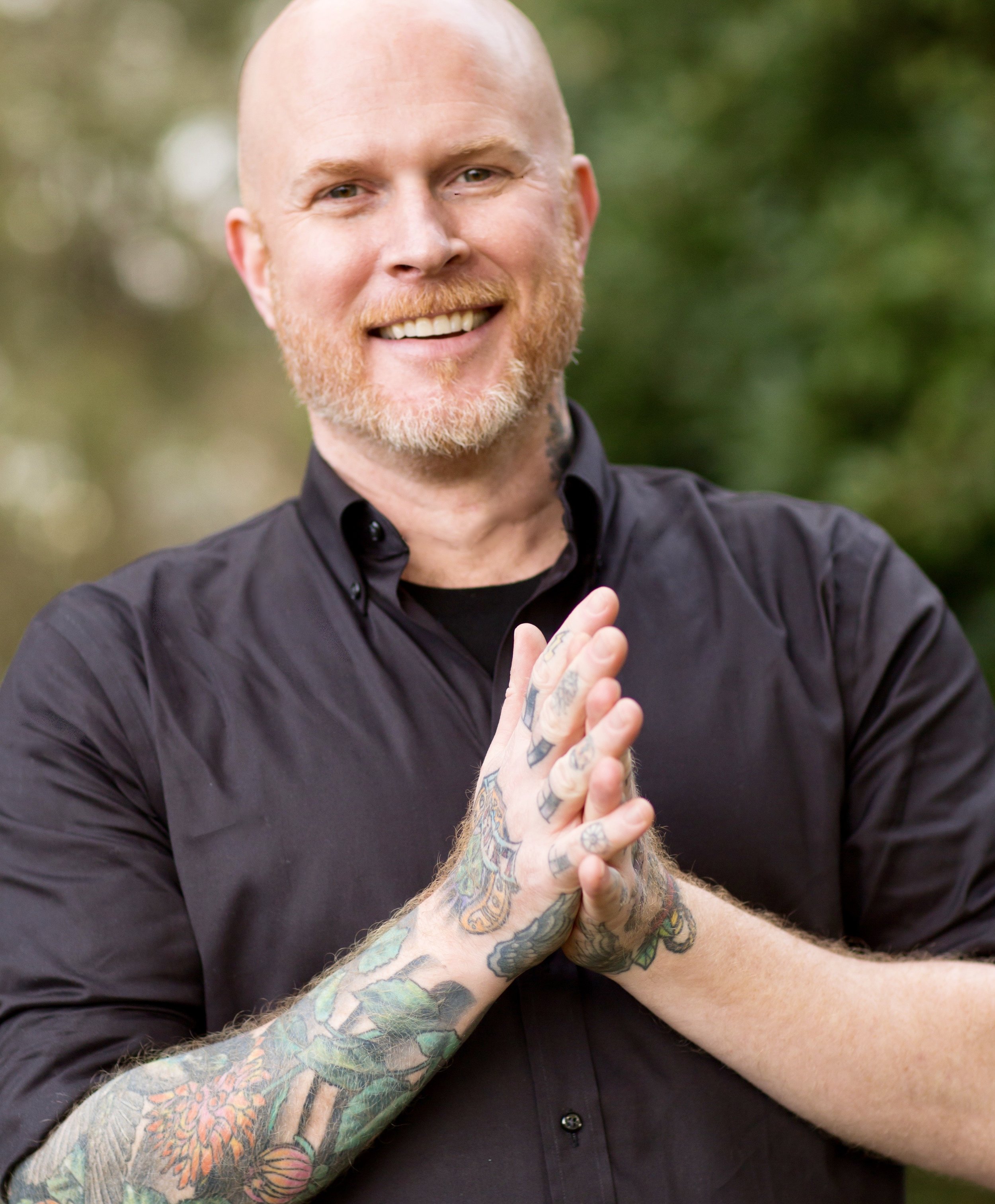 A smiling man with a bald head, beard, and tattoos on his arms, wearing a black shirt, outdoors with blurred green foliage background.