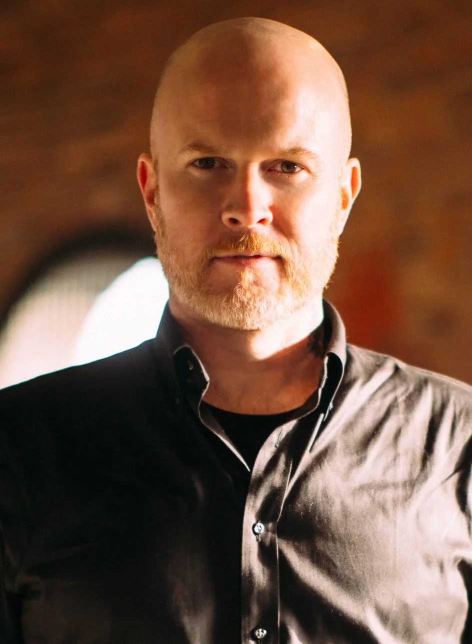 A close-up portrait of a bald man with a beard, wearing a black shirt, against a blurred background.