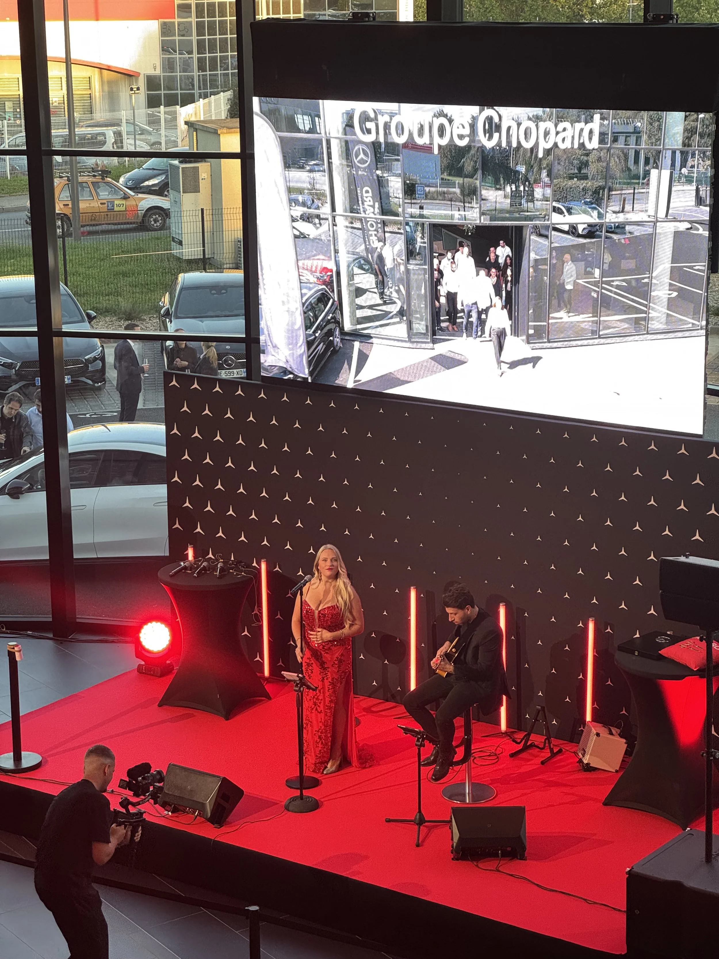 Une scène d'événement avec une chanteuse en robe rouge et un musicien jouant de la guitare sur scène rouge, une grande affiche avec le logo Mercedes et Chopard en arrière-plan. Des personnes regardent et prennent des photos, avec une grande fenêtre derrière apportant de la lumière naturelle.