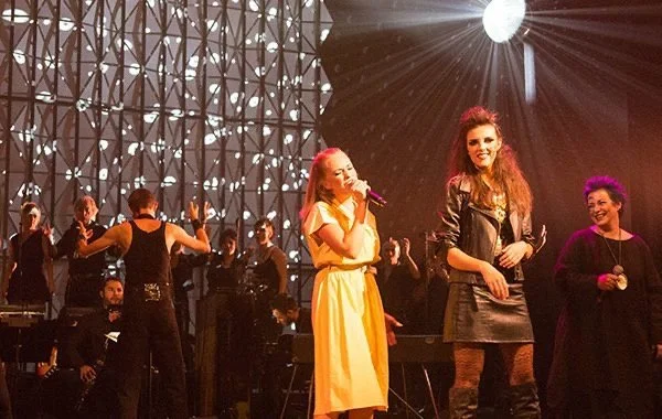 Trois femmes chantent et dansent lors d'une performance musicale sur scène, avec un orchestre en arrière-plan et un éclairage dramatique.