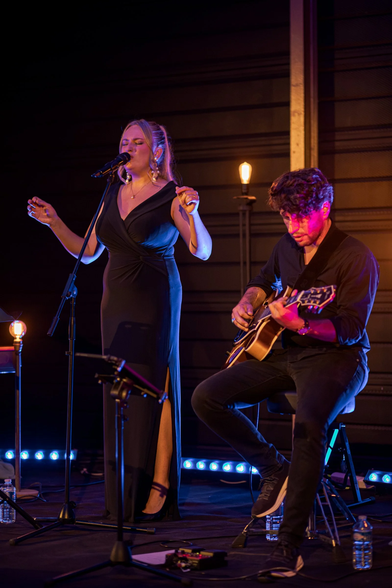 Une femme chante avec un homme jouant de la guitare acoustique lors d'une performance musicale dans une ambiance chaleureuse, avec des lampes et une scène en arrière-plan.