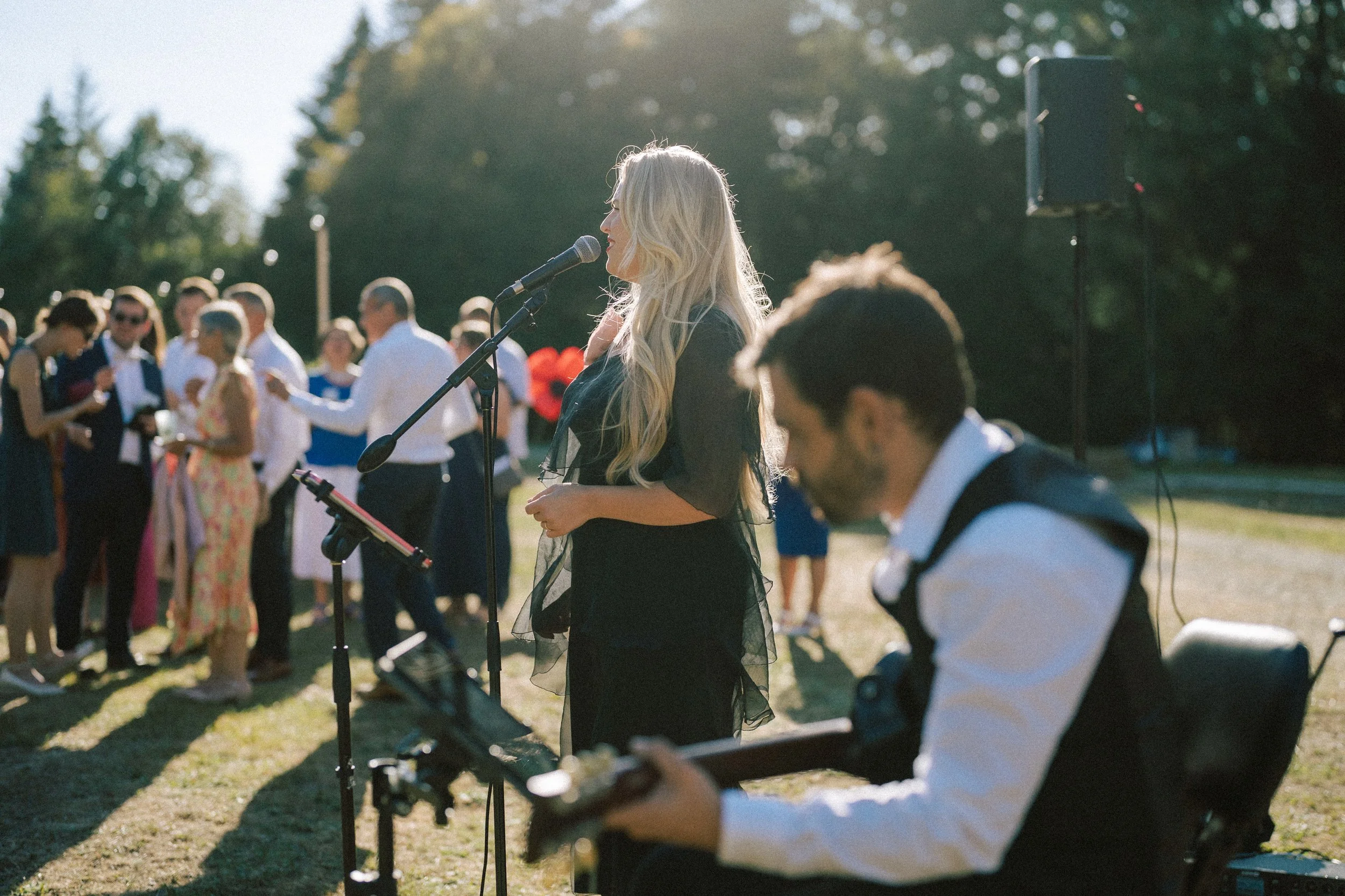 Une femme blonde chante dans un microphone lors d'un événement en plein air, entourée d'autres personnes qui dansent et jouent de la musique sous un soleil d'après-midi.