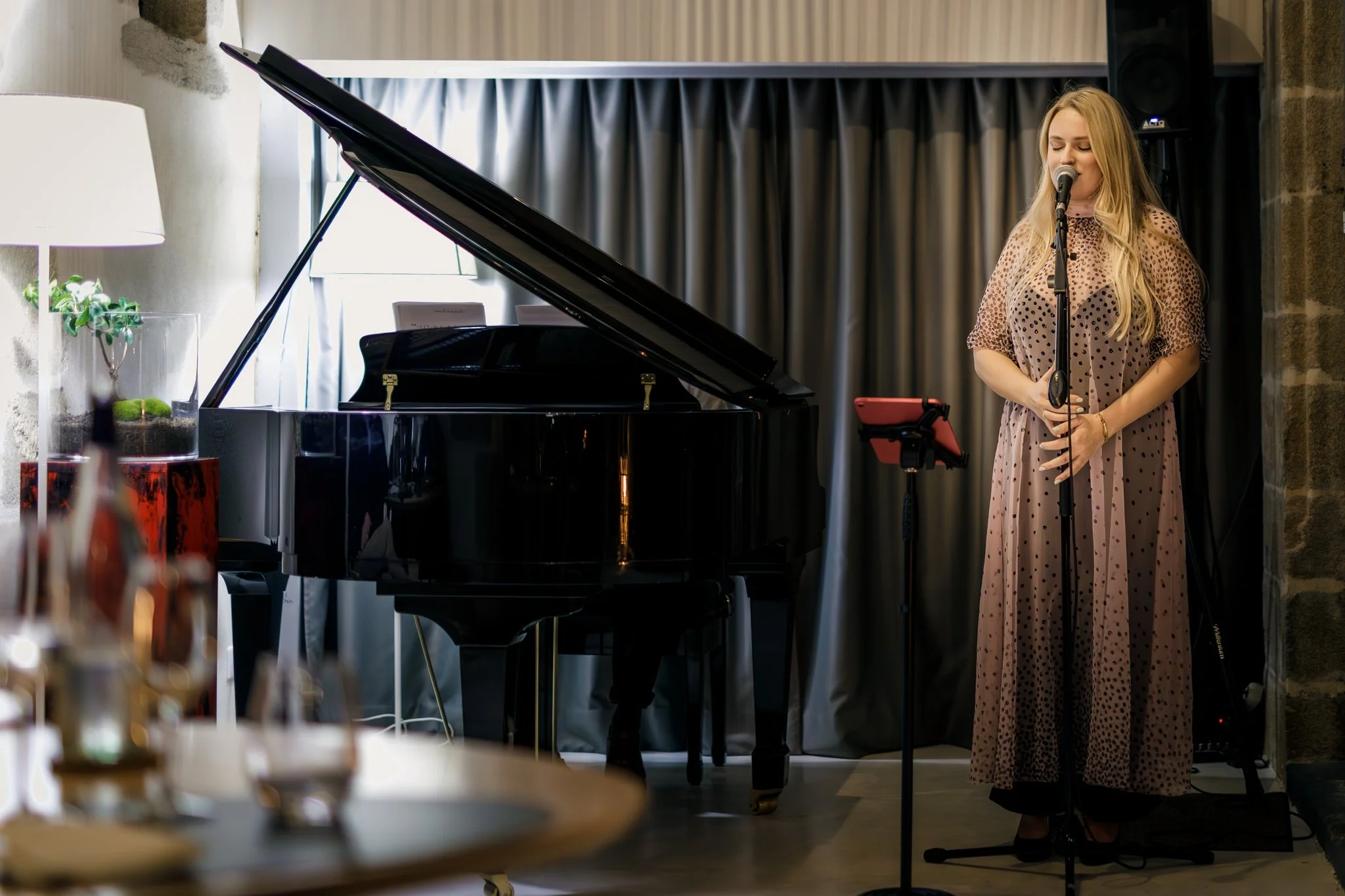 Une femme chante devant un micro, à côté d'un piano noir, dans une pièce bien éclairée avec un rideau sombre en arrière-plan.