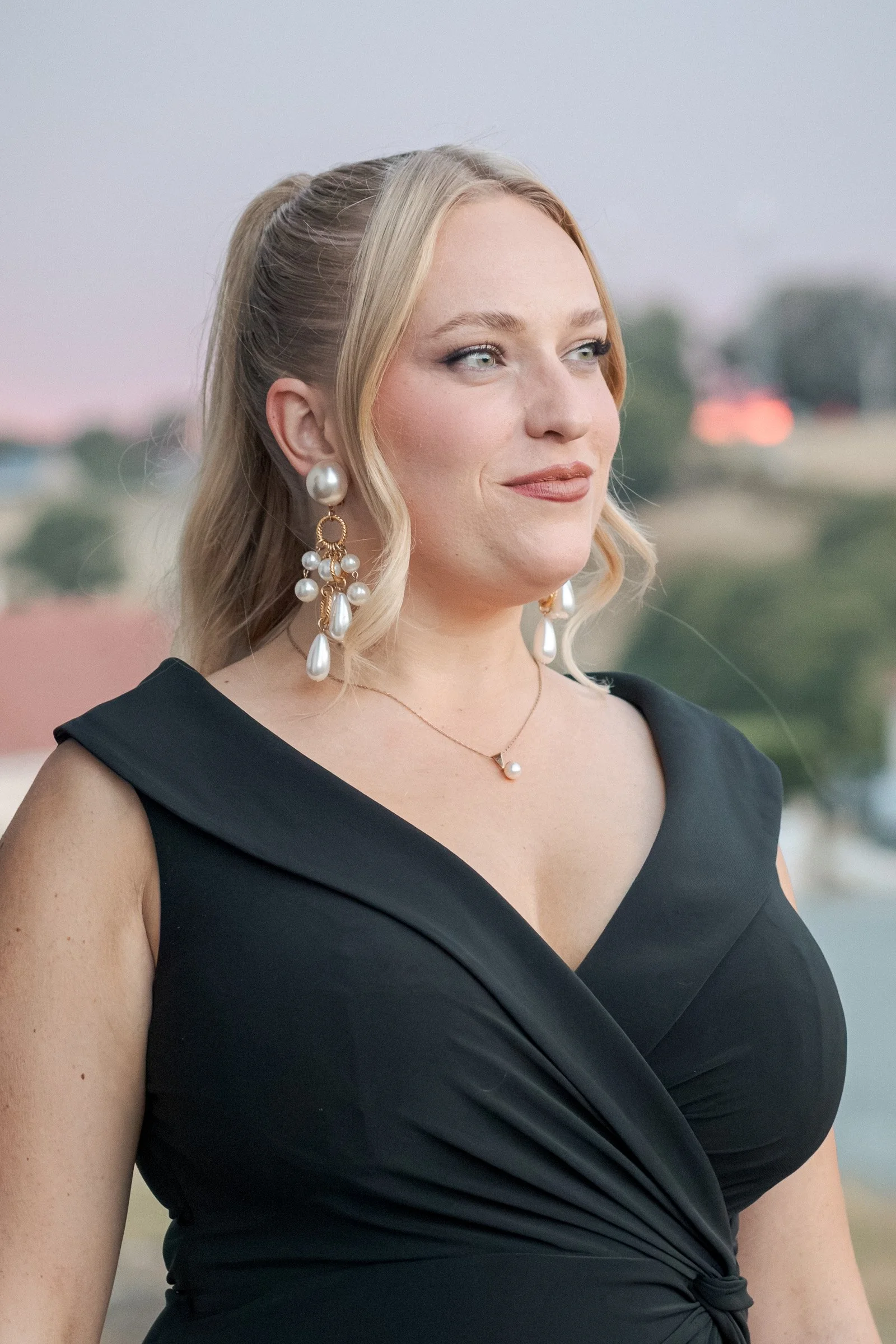 Une femme porte une robe noire avec un décolleté en V et des boucles d'oreilles pendantes en perles, avec un collier en perle. Elle a des cheveux blonds attachés en un chignon bas avec quelques mèches lâches autour du visage, sourit légèrement en regardant vers la droite. Le fond est flou avec un ciel au crépuscule et des arrières-plans urbains.