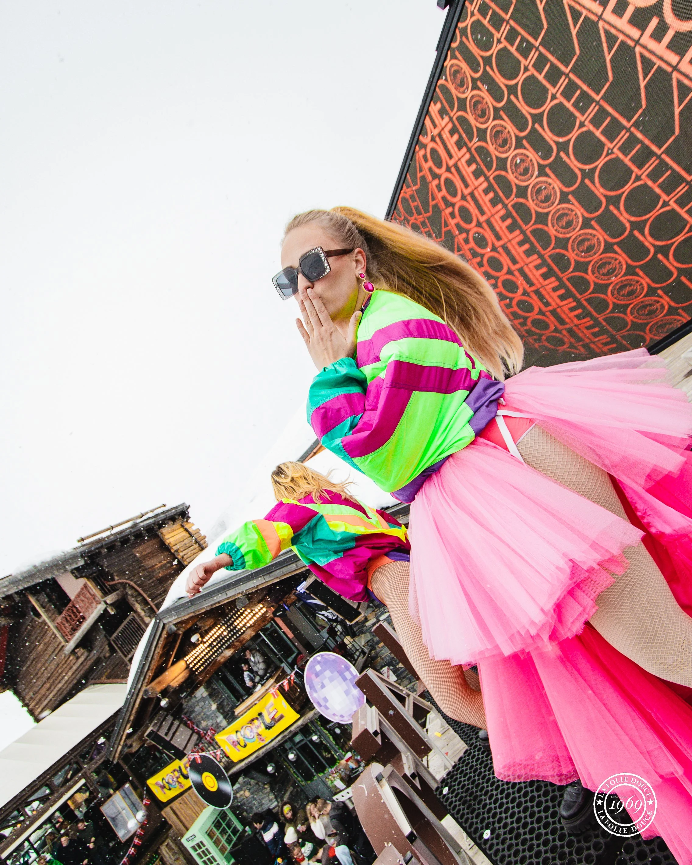 Une femme avec des vêtements colorés et une jupe en tulle rose, portant de grosses lunettes de soleil et des boucles d'oreilles roses, pose devant un mur de publicité avec le mot 'Foile' répété, dans un lieu public animé.