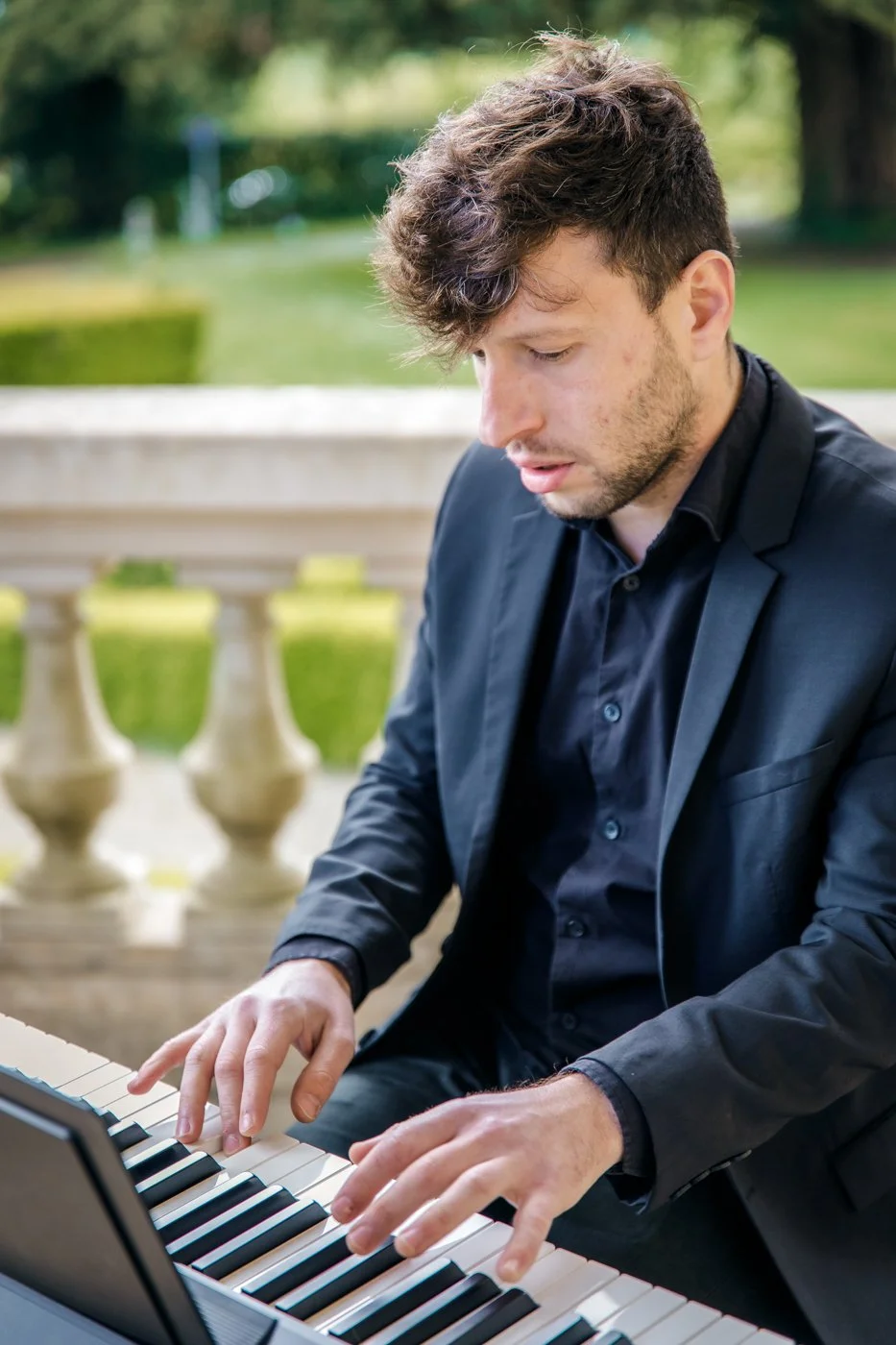 Jeune homme jouant du piano en extérieur, portant un costume noir, avec un fond de nature verdoyante.