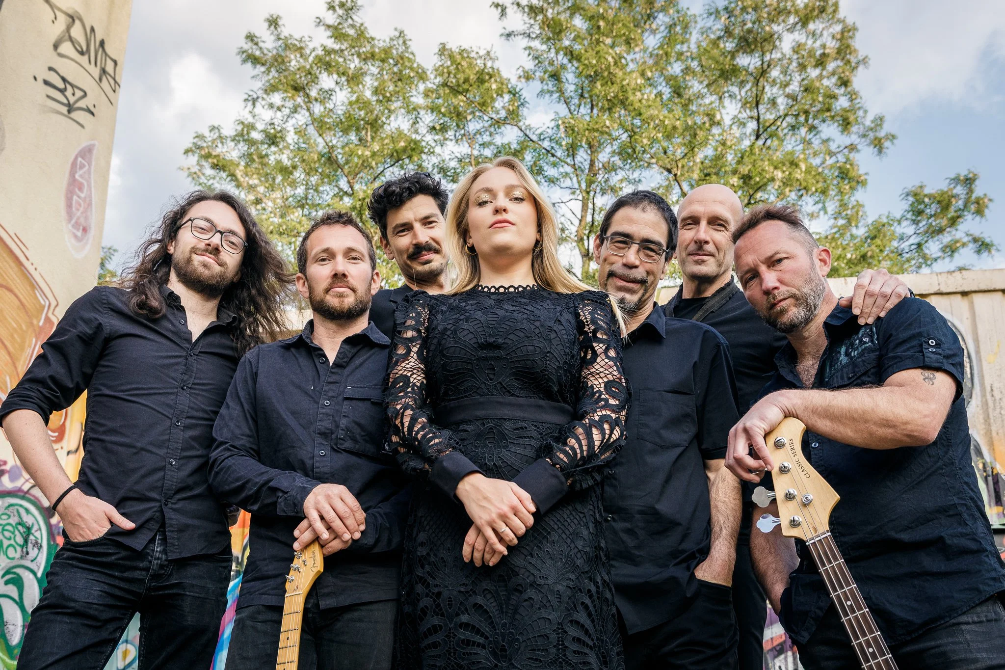 Groupe de sept musiciens, dont une femme, posant devant un mur graffité et des arbres, avec deux guitares, lors d'un événement en plein air.