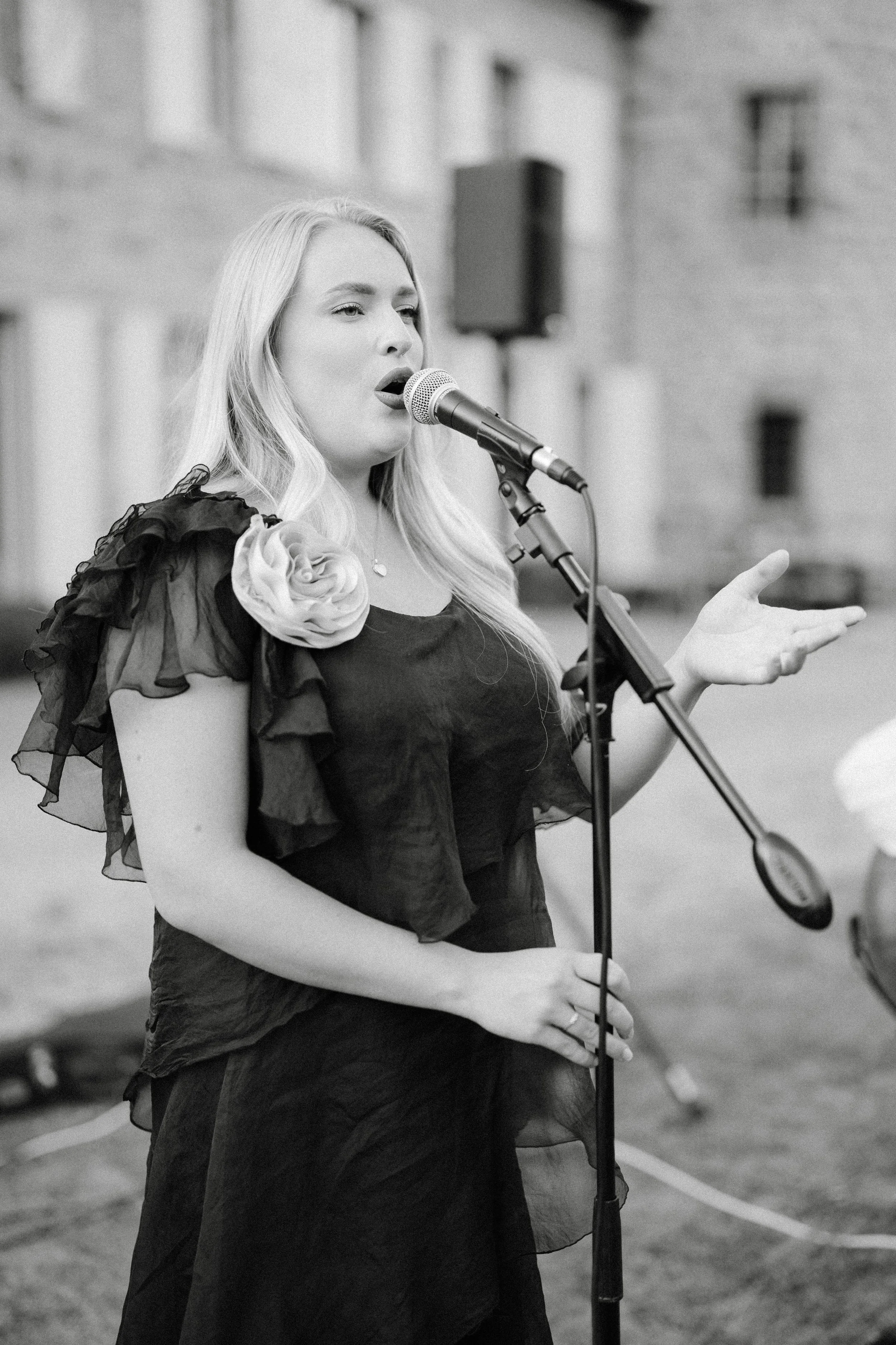 Une femme blonde chante ou parle dans un micro, vêtue d'une robe noire avec des détails en tissu transparent, décorée d'une grande rose en tissu sur son épaule, lors d'un événement en extérieur.