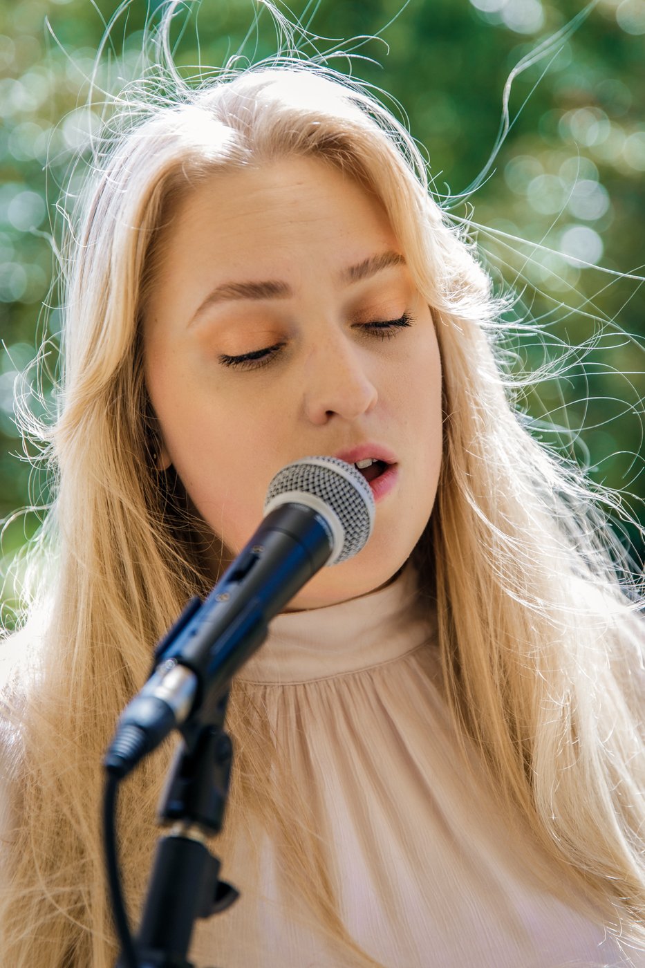 Jeune femme blonde chantant dans un microphone en plein air avec un fond flou de verdure.