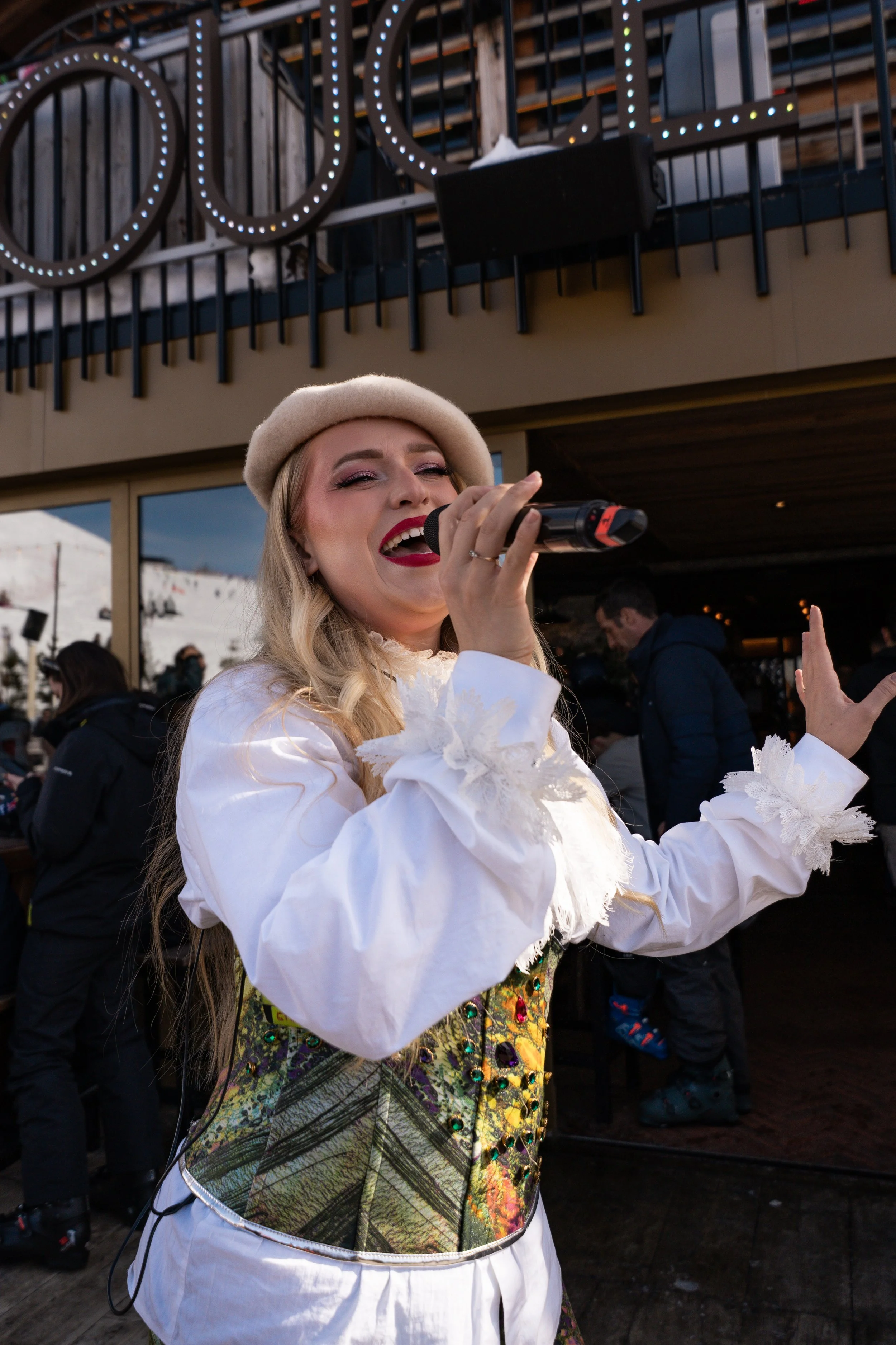 Femme souriante portant un chapeau beige et une tenue colorée, chantant avec un microphone lors d'un événement extérieur en montagne.